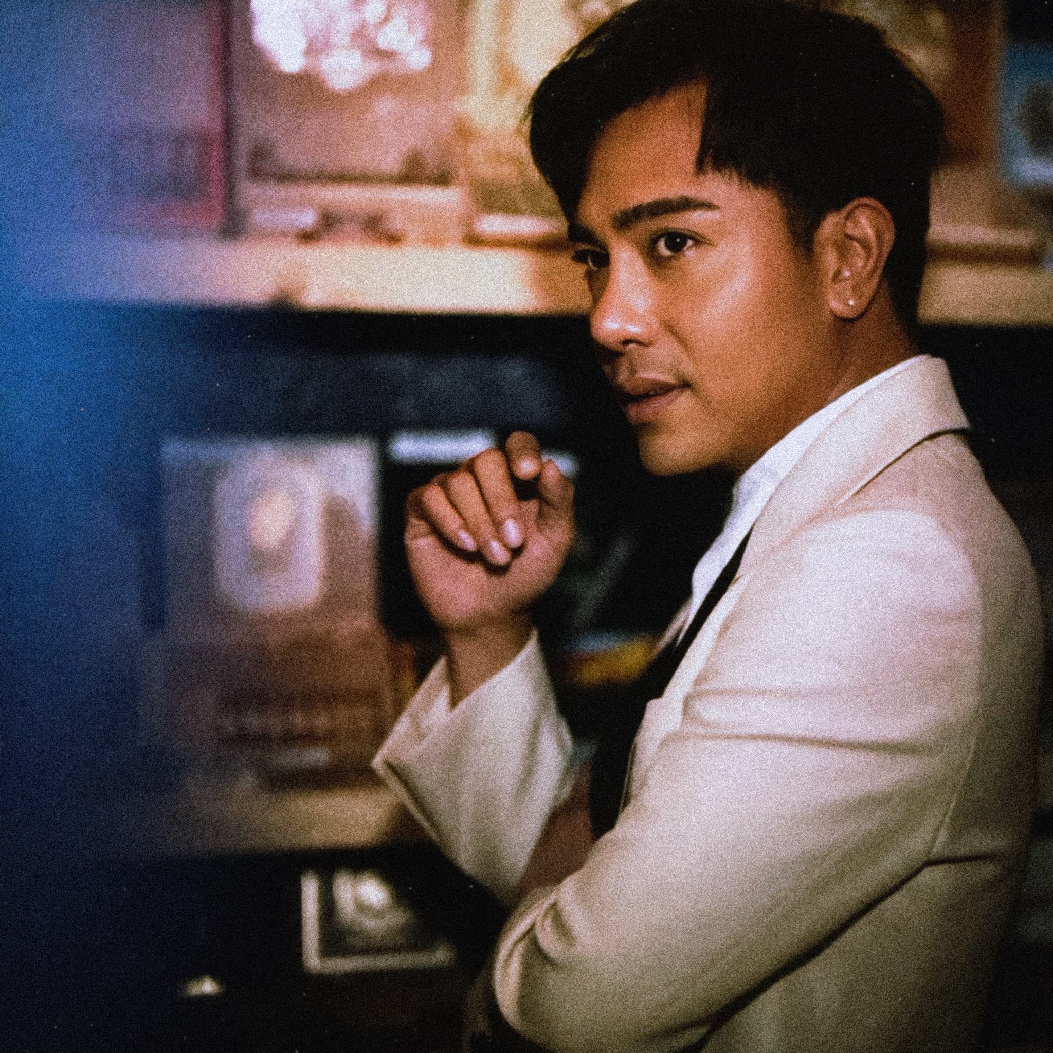 Close portrait of Raffy Bleu in a white dinner jacket, hands clasped at his chin, looking to camera across a lit shop counter